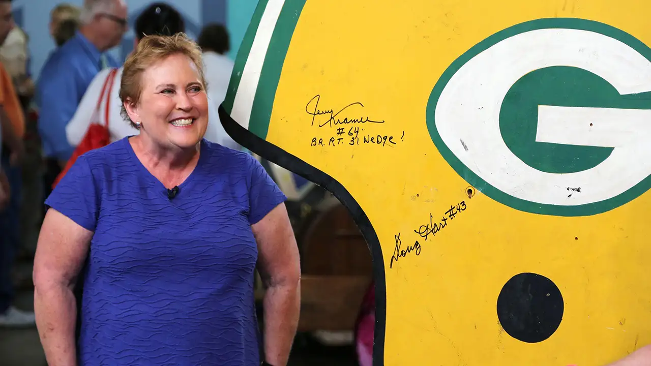A smiling woman in a blue shirt stands beside a large yellow and green sign with autographs, including "Jerry Kramer #64 BR RT 31 Wedge" and "Doug Hart #43".