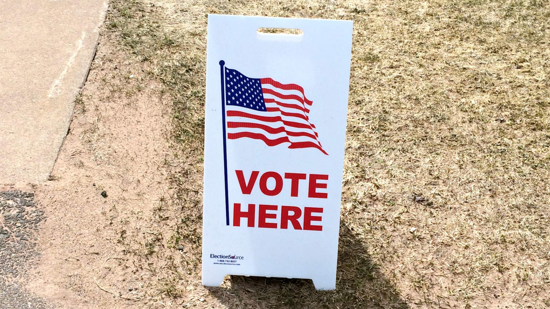 A sandwich board sign that says "Vote Here" with an American flag above it