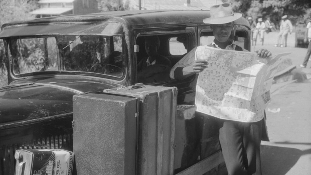 An African American man reads a road map while leaning against a car.
