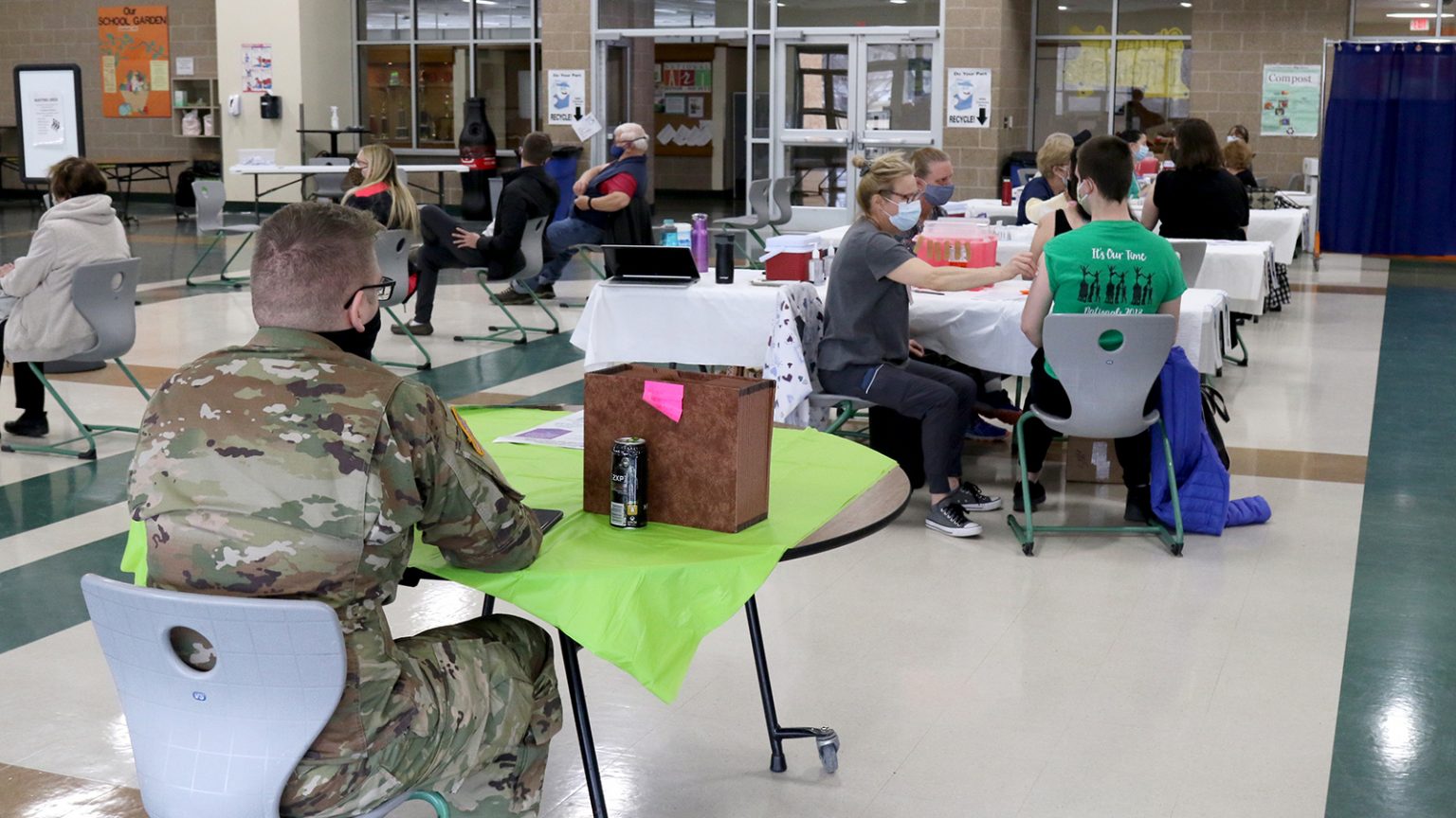 A Wisconsin National Guard member sits at a table at as vaccinators give coronavirus vaccines to patients at other tables.