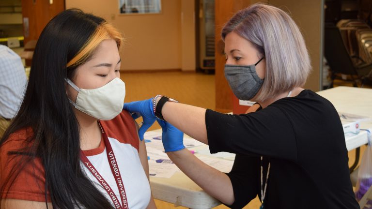 A vaccinator wearing gloves and mask gives a shot to another person wearing a mask.