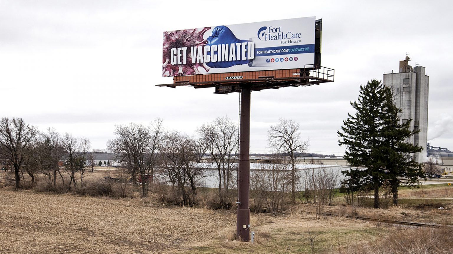 A billboard reading Get Vaccinated showing a boxing glove punching a coronavirus viroid illustration is surrounded by farm fields and warehouses.