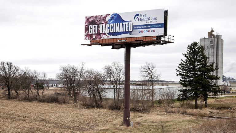 A billboard reading Get Vaccinated showing a boxing glove punching a coronavirus viroid illustration is surrounded by farm fields and warehouses.