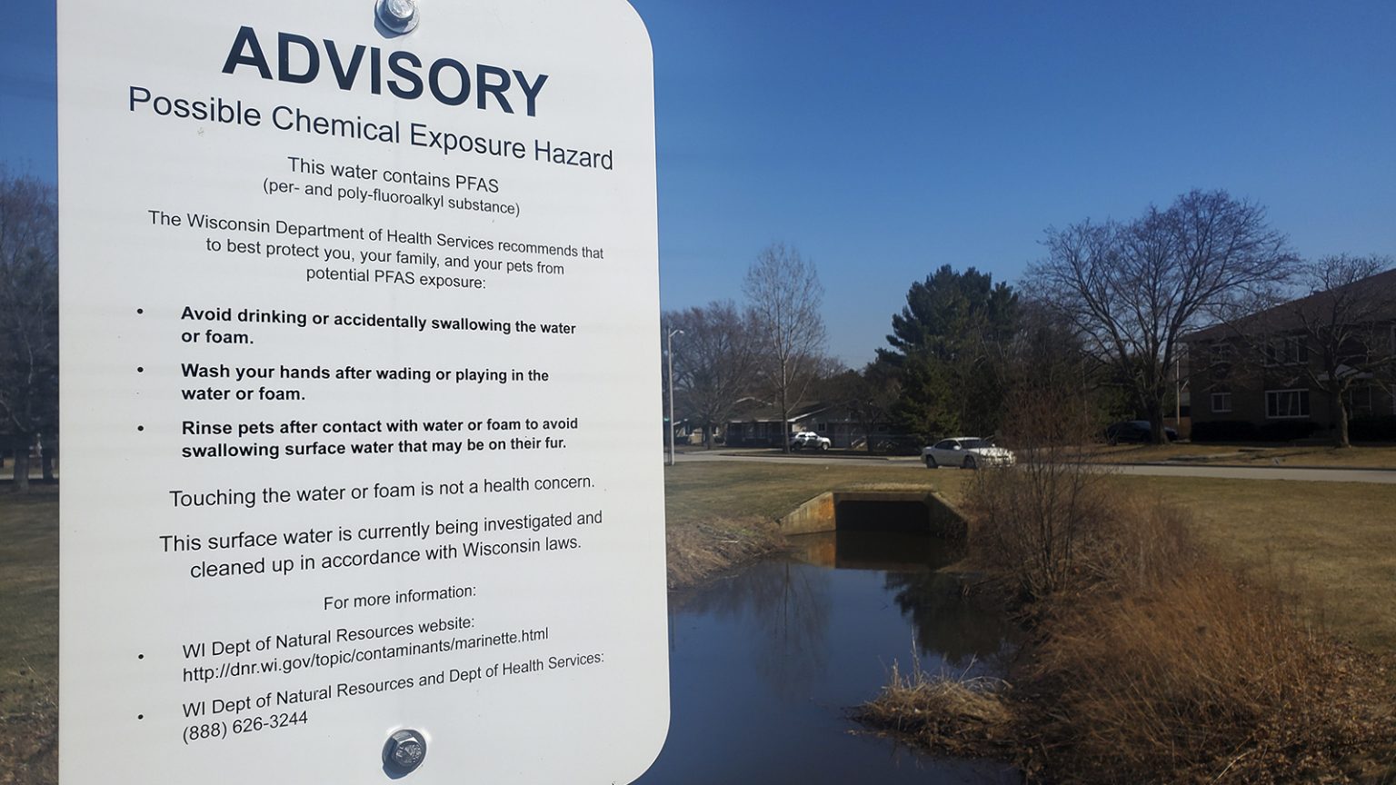 Sign warning of possible chemical exposure hazard above ditch and culvert with houses in background