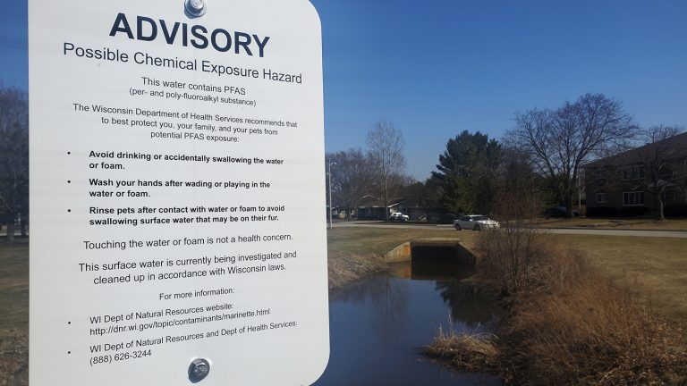 Sign warning of possible chemical exposure hazard above ditch and culvert with houses in background