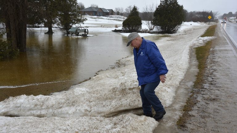 Person standing in snowbank and collects water sample from flooded yard.