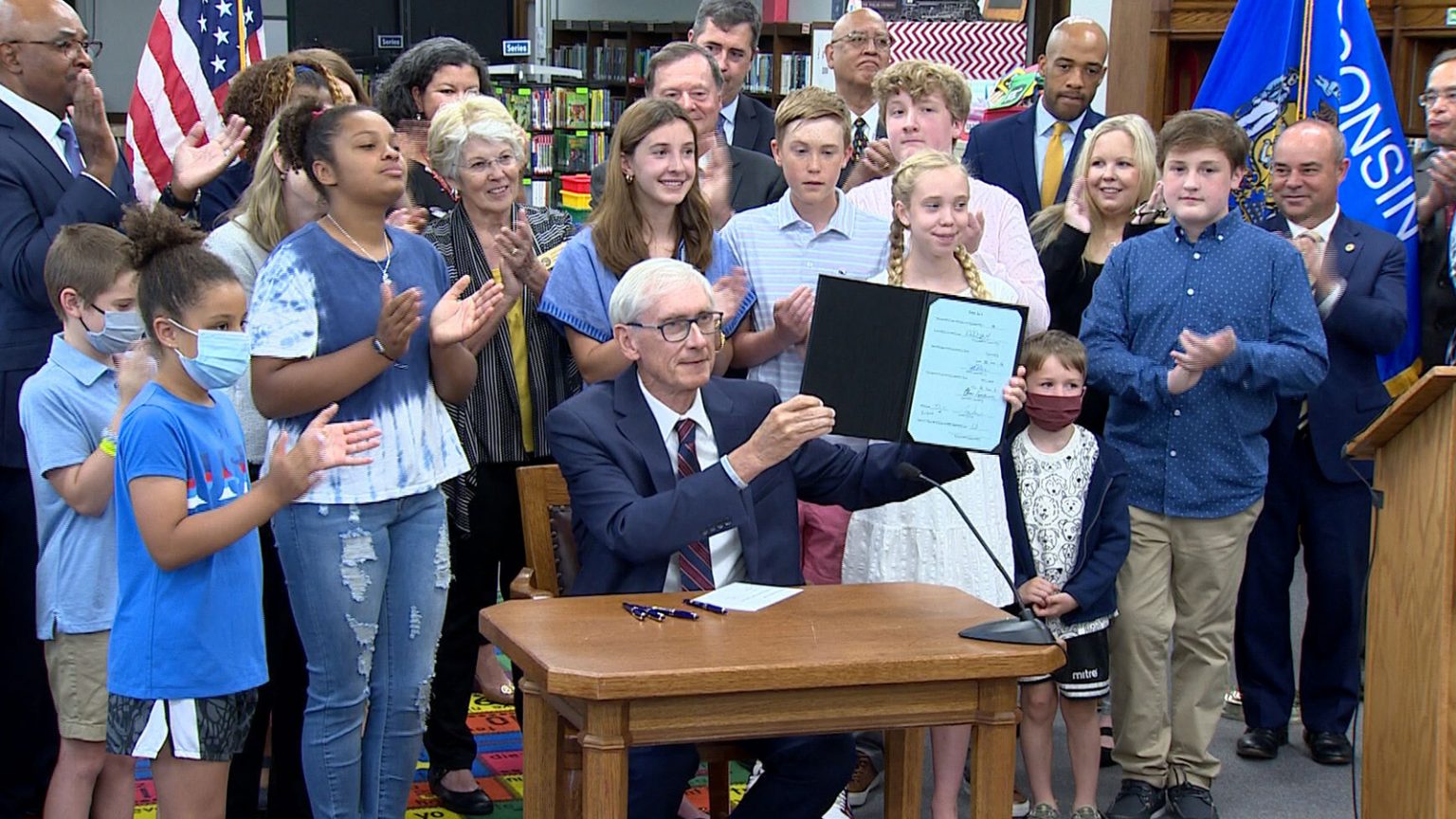 Tony Evers sitting at table in a classroom holding budget document surrounded by state officials and school children