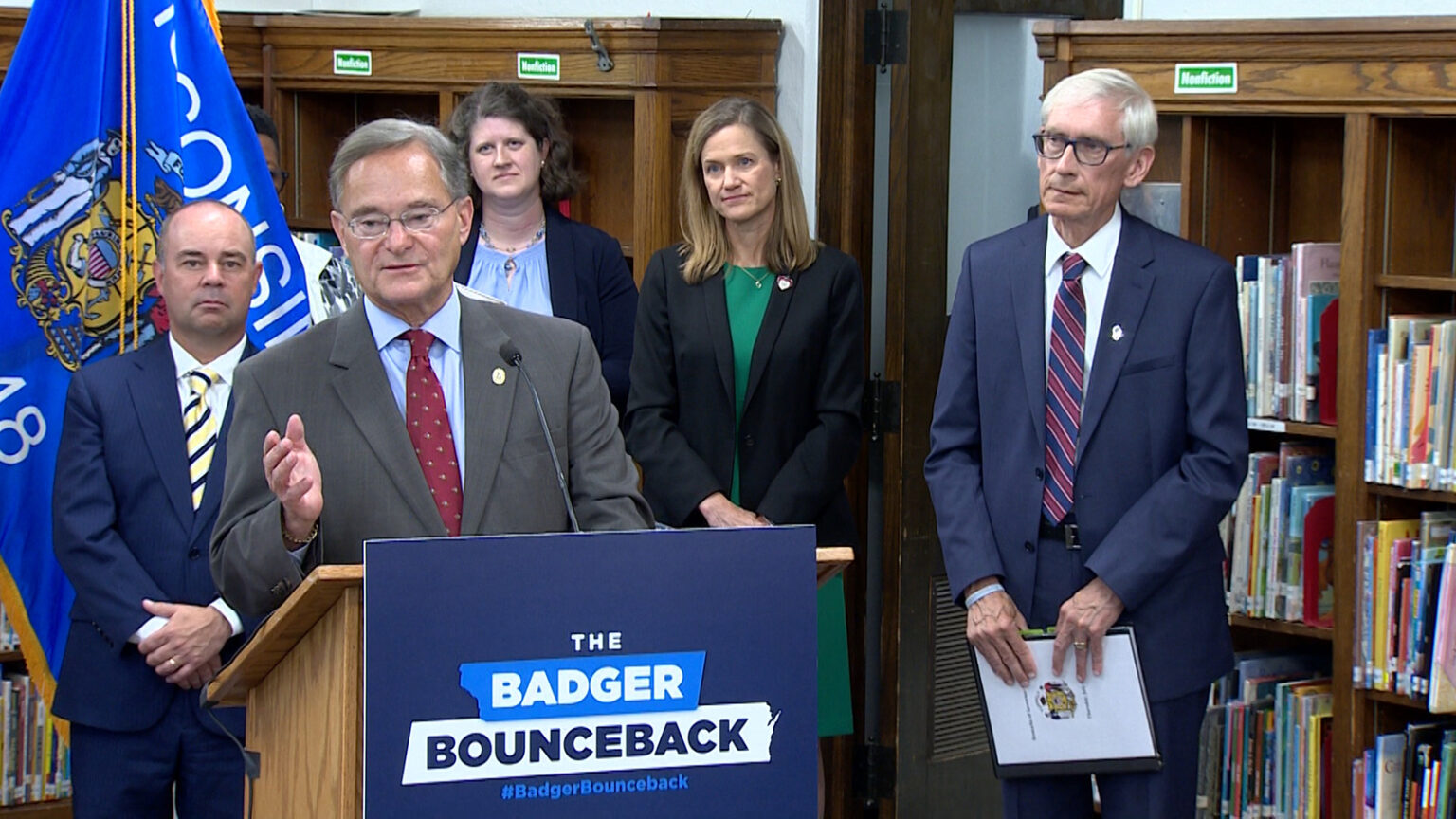 Peter Barca standing at podium in school library surrounded by Tony Evers and other members of the governor's administration