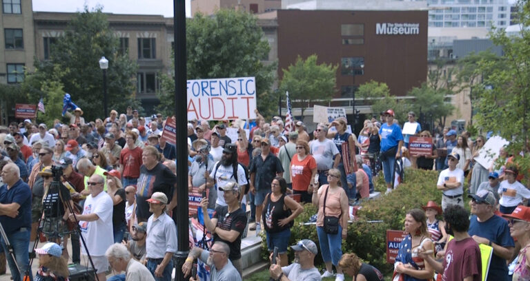 Protestors gather in front of the State Street entrance of th Wisconsin Capitol, with one holding a sign reading Forensic Audit. 