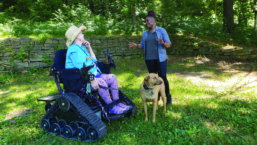 A woman in a wheelchair holds her dog on a leash. She talks to a woman standing next to her.