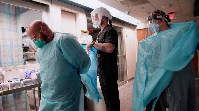 Three hospital workers put on personal protective equipment including gowns, masks and face shields in the hallway of an intensive care unit.
