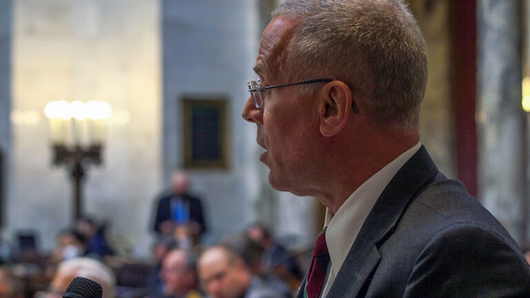 A profile portrait of Rep. Chuck Wichgers on the Wisconsin Assembly floor with seated legislators in the background.