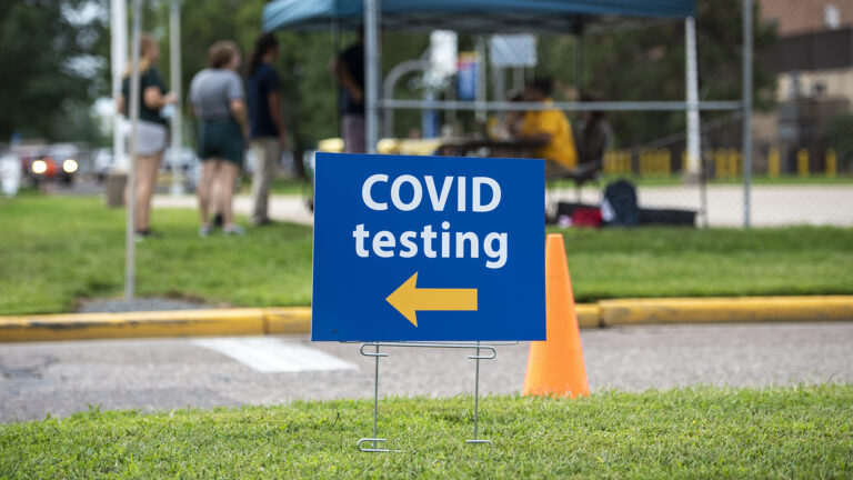 A yard sign stating COVID testing with an arrow pointing left stands in front of a canopy with a table underneath and a queue of people in front of a table.