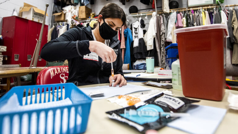 A nurse prepares a syringe in a storage room with theater costumes hanging in the background.