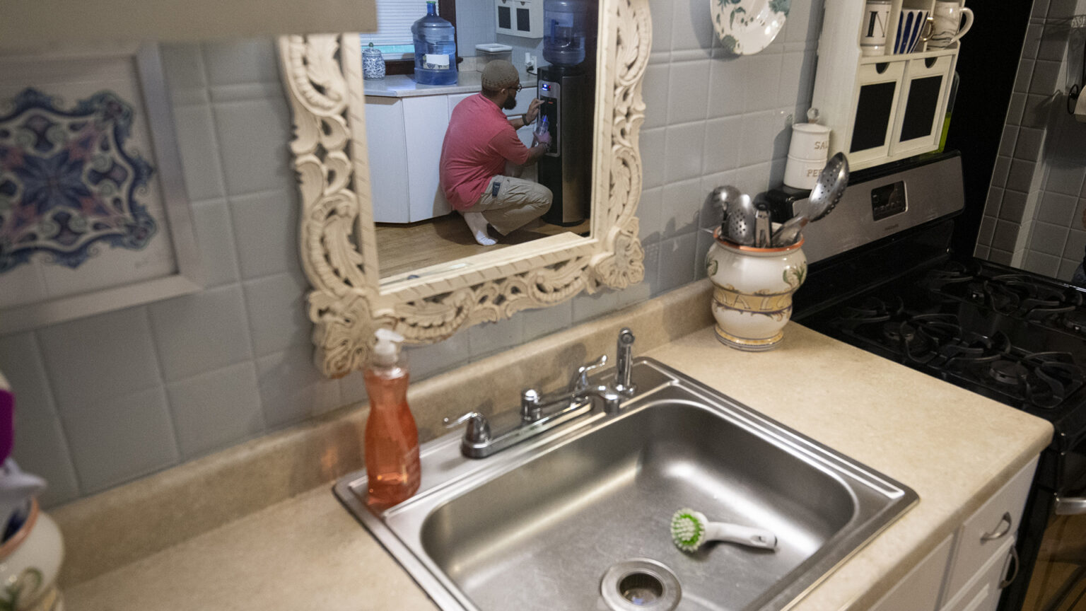 Nazir Al-Mujaahid fills a water bottle from a water system as seen in a mirror above a kitchen sink.