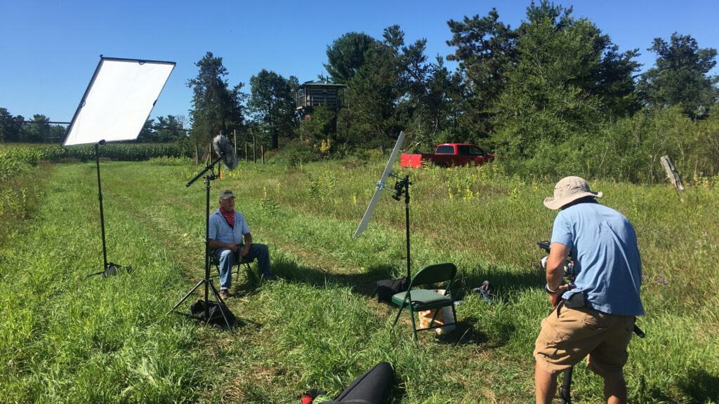 A farmer is interviewed by a camera crew in front of a crop field