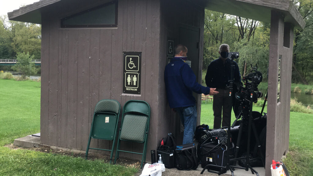 Camera man and interviewee take shelter from the rain next to an outdoor bathroom