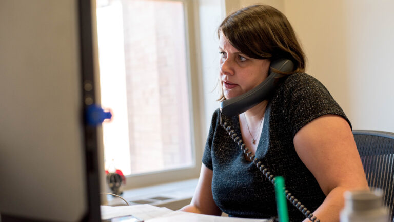 Claire Woodall-Vogg sits at a desk with an office phone resting betweeen her ear and shoulder.