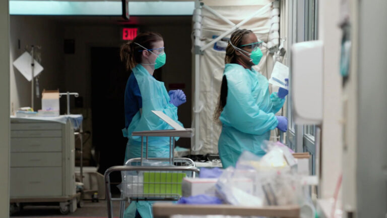 Two people wear medical gowns, gloves, masks and goggles as they prepare to enter a room inside a hallway filled with medical equipment