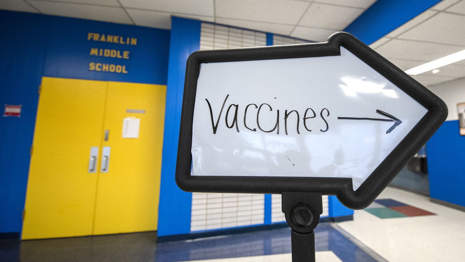 A whiteboard in the shape of an arrow with Vaccines and an arrow written on it stands in a school hallway.