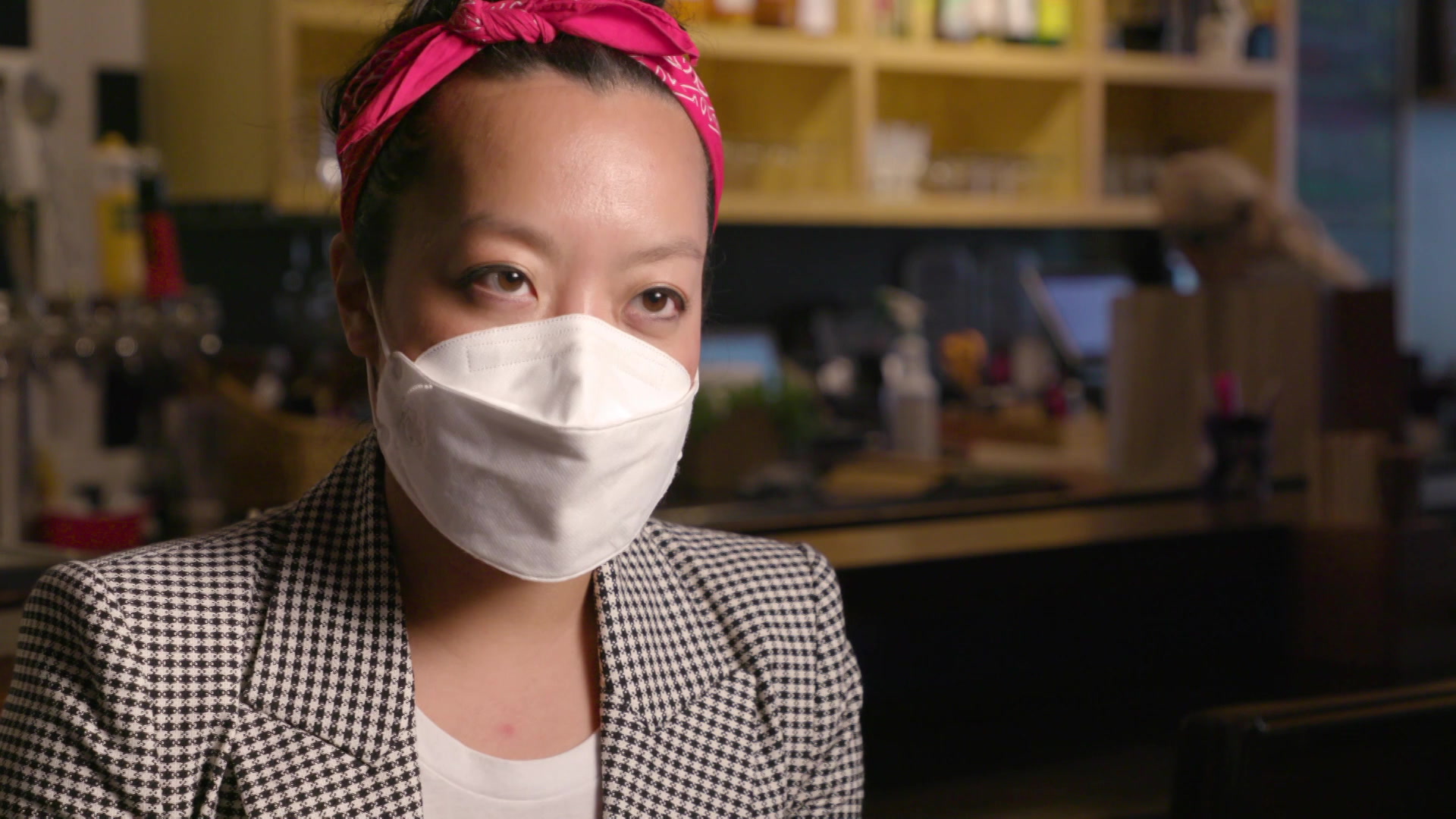 A portrait of Francesca Hong, wearing a suit and face mask with beer taps and cabinets in the background.