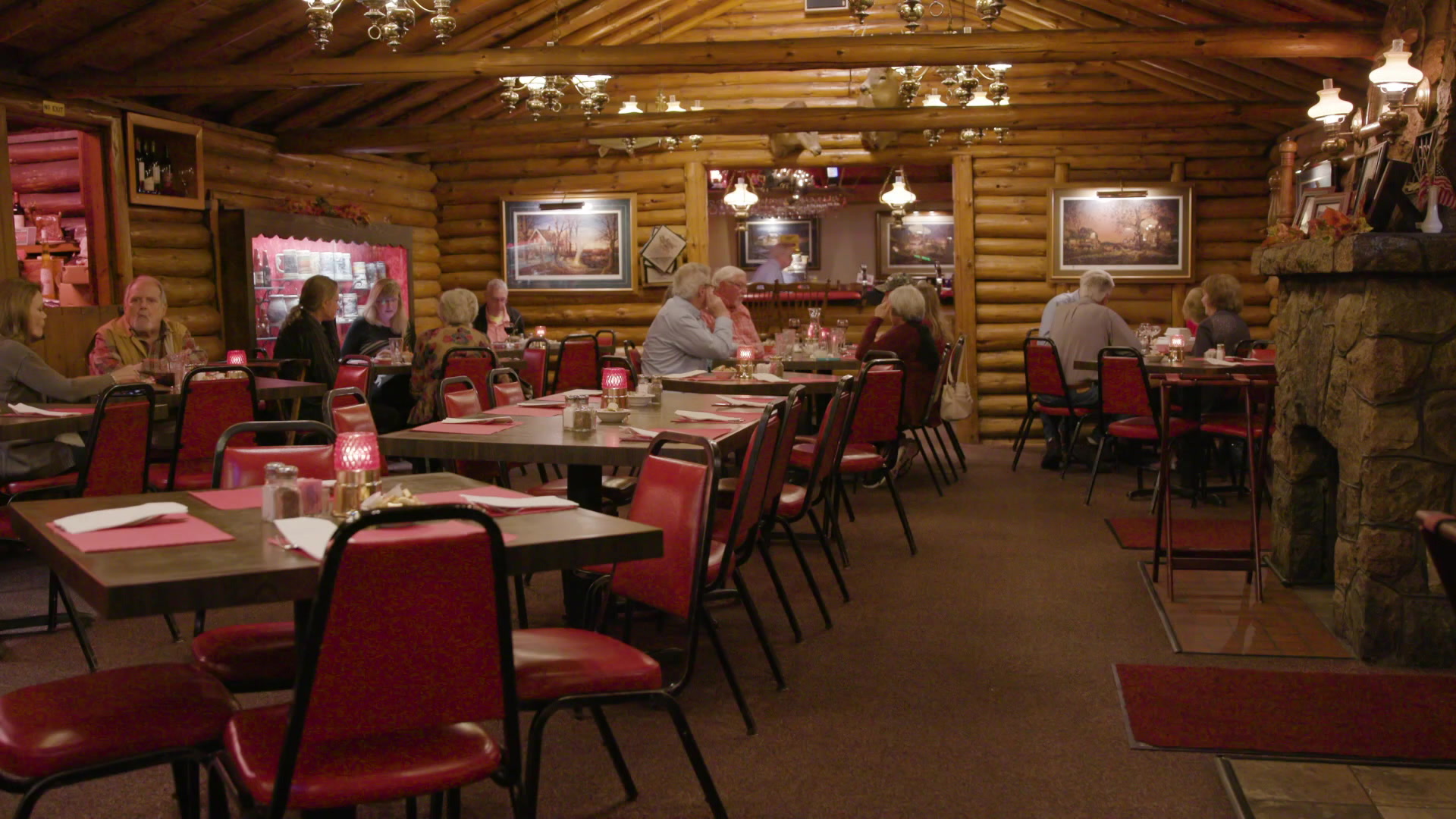 Dinners sit on red chairs and at tables inside a dining room with log walls and a stone fireplace at right.