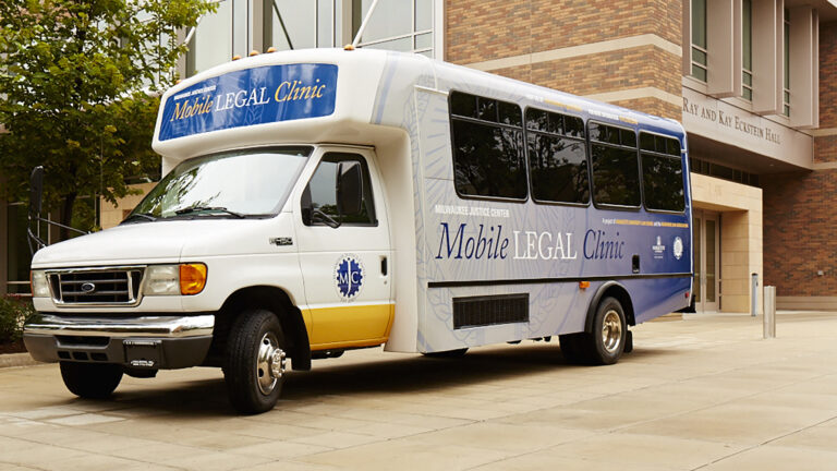 A trailer-style bus with signage stating Milwaukee Justice Center Mobile Legal Clinic is parked in front of a building.