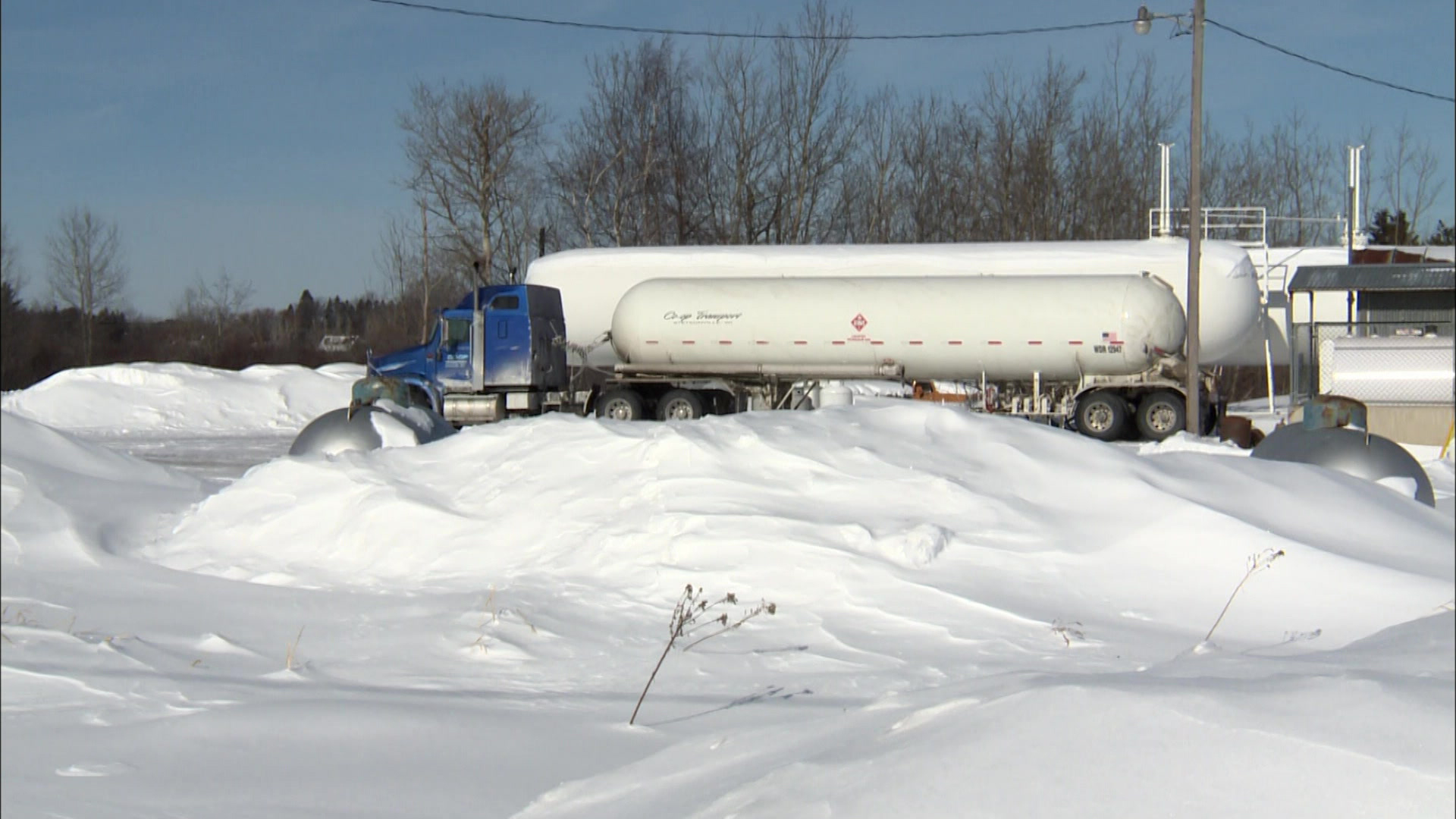 A propane truck sits parked in front of larger propane storage tanks, with smaller propane storage tanks in the foreground mostly covered by snowbanks.