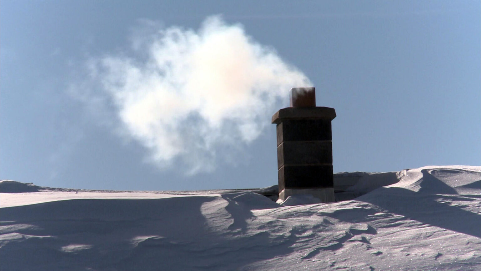 Smoke flows out of of a masonry chimney on a snow-covered roof.