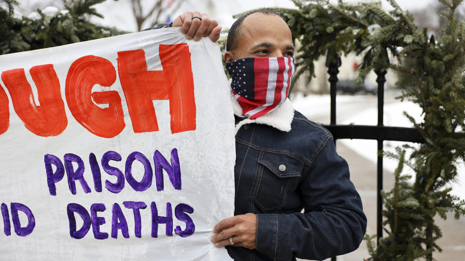 Ramiah Whiteside wears an American flag mask and holds a protest sign in front of a wrought iron fence.