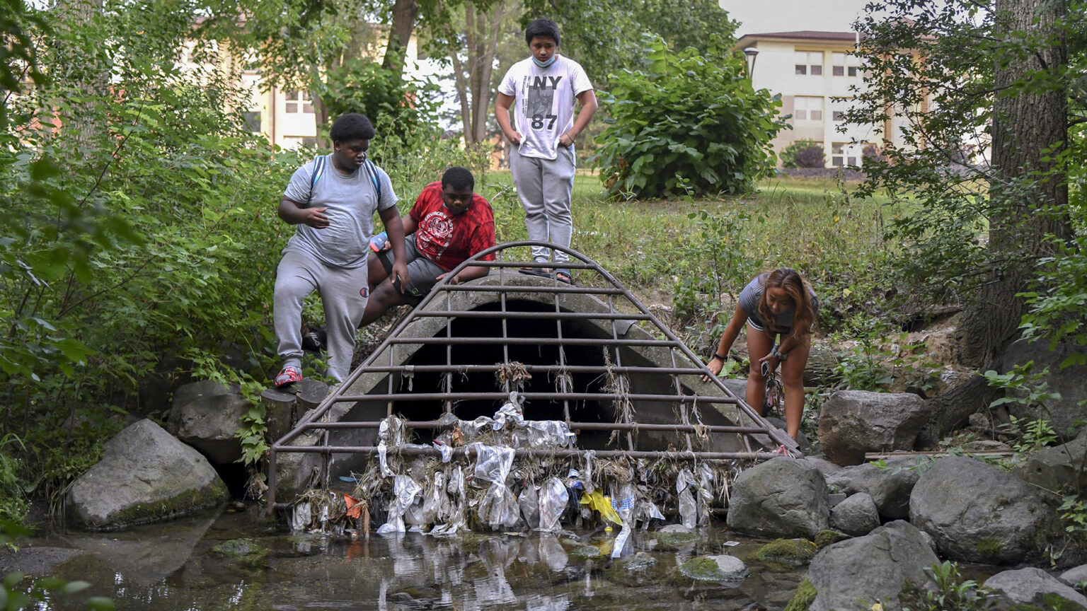 Four children stand on a concrete culvert mouth with a metal grille covered with plastic garbage on the edge of a creek.