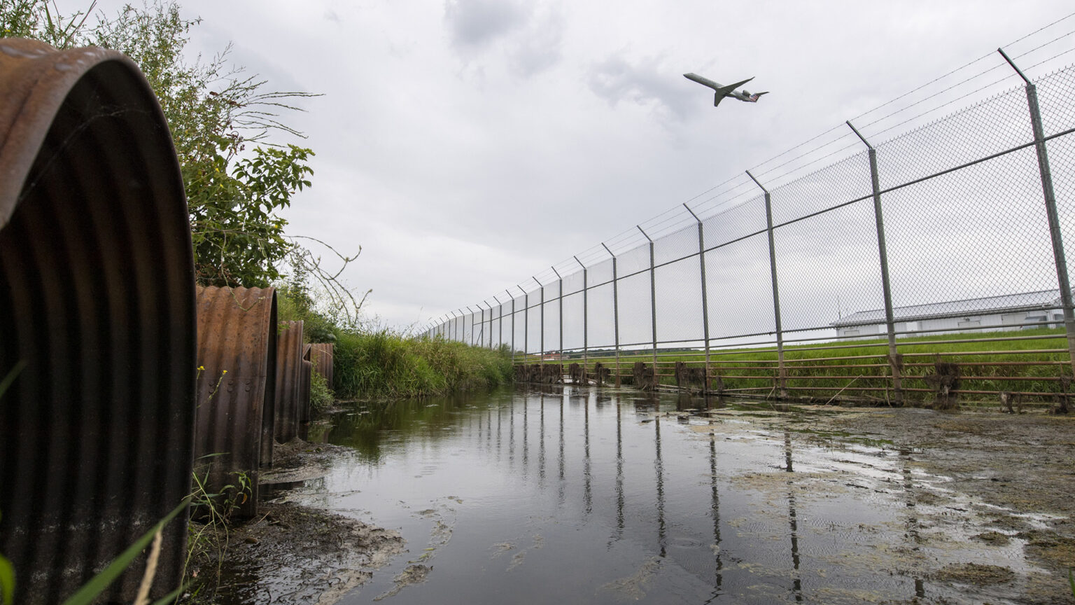 An airplane ascends over a chain-linked fence topped with barbed wire that faces a wetland area with rusty culvert mouths.