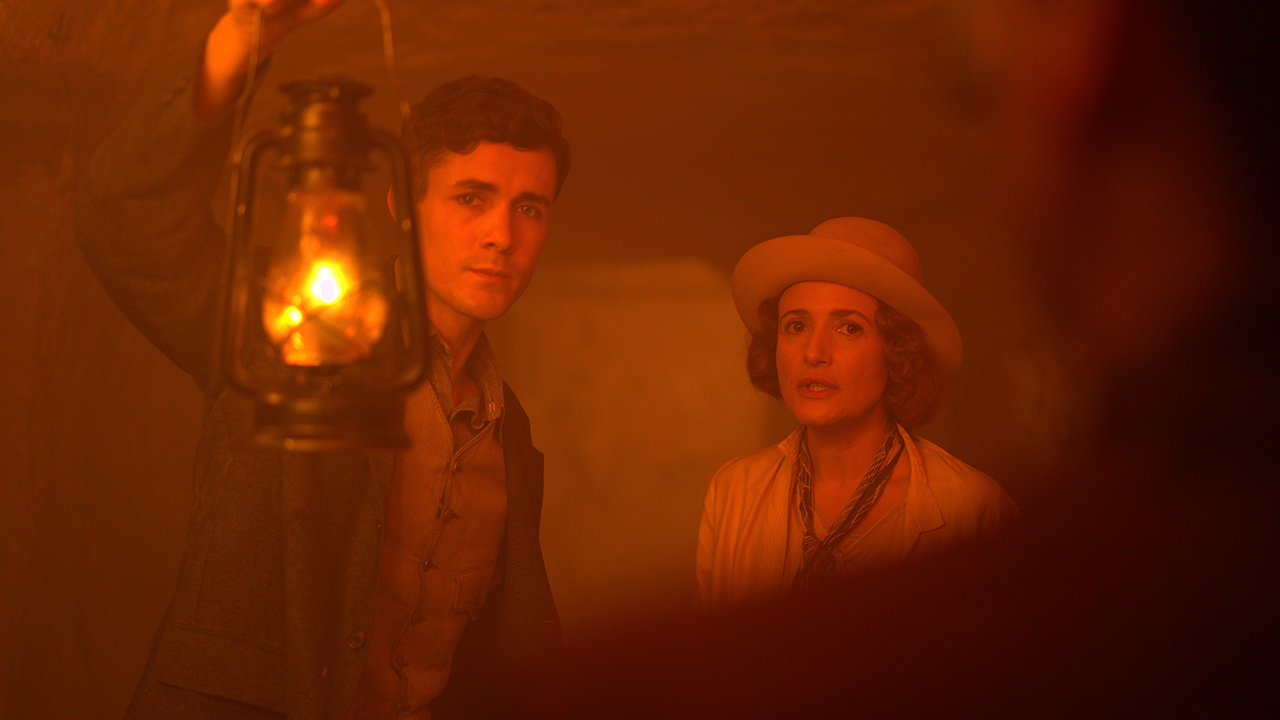 A man holds up a kerosene lantern toward the camera. A woman in a hat stands next to him.
