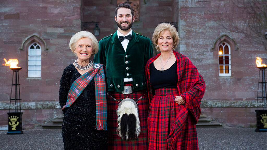 Two women and a man dressed in holiday attire stand before a house.