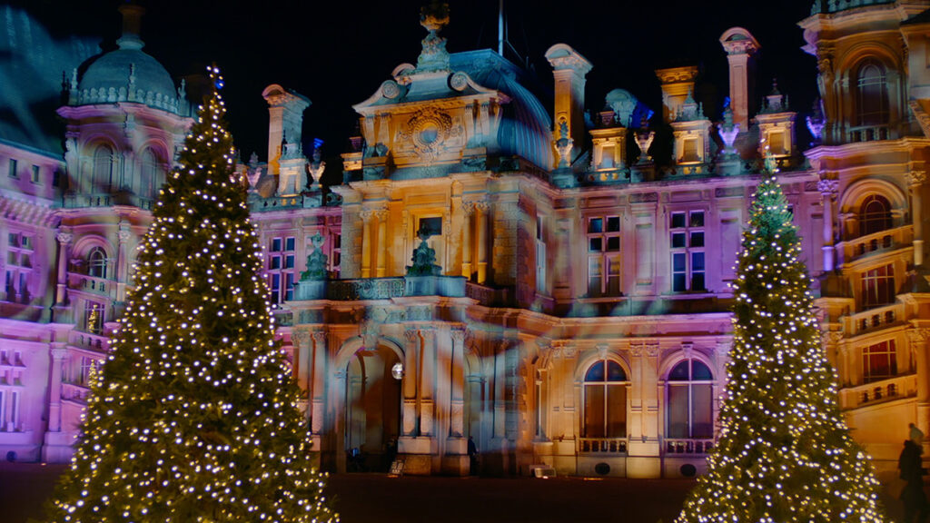 A nighttime photograph of a Christmas tree lit up. A large house is in the background with colorful lights shining on the front of it.