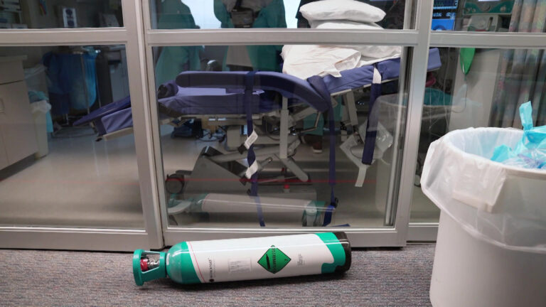 A discarded oxygen tank sits on the floor outside the entrance of a hospital ICU room with medical personnel standing next to an empty bed stacked with pillows.