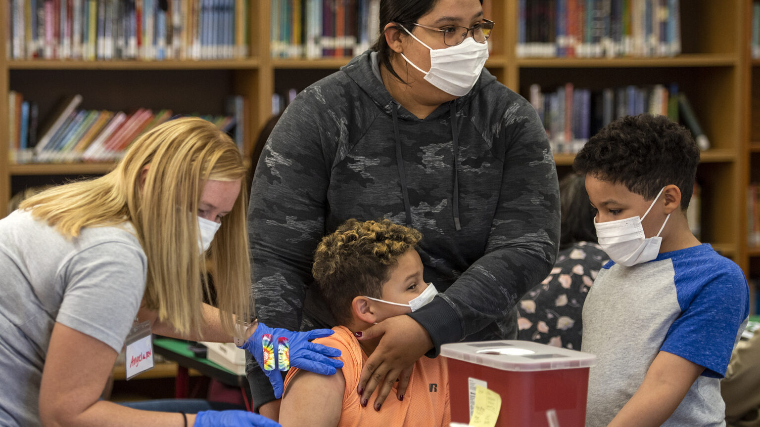 Inside a library, a seated child is held by an adult as another adult administers a shot and a second child watches.