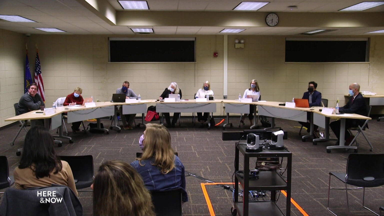 School board members sit at tables arranged in a semi-circle in front of seated audience members and a cart holding audio-visual equipment.