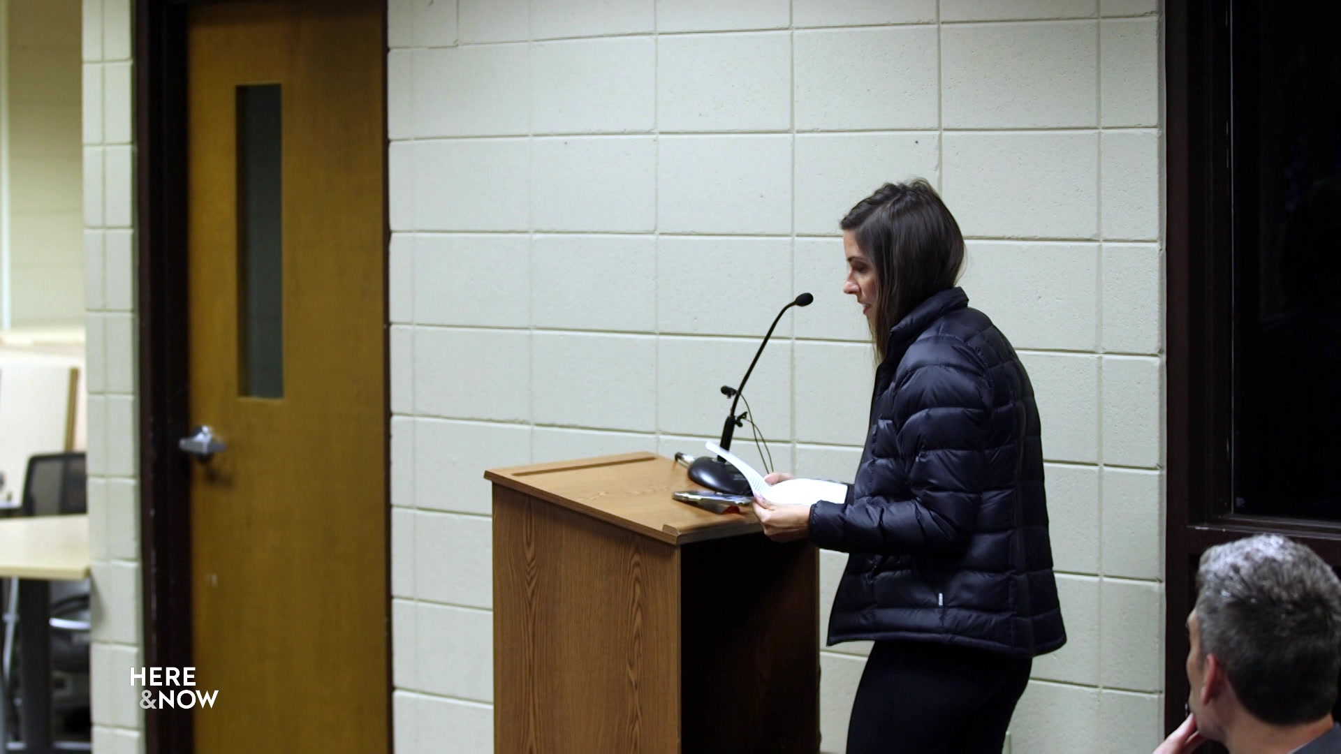 Amber Schroeder stands at a podium holding a piece of paper in front of a painted concrete-block wall.