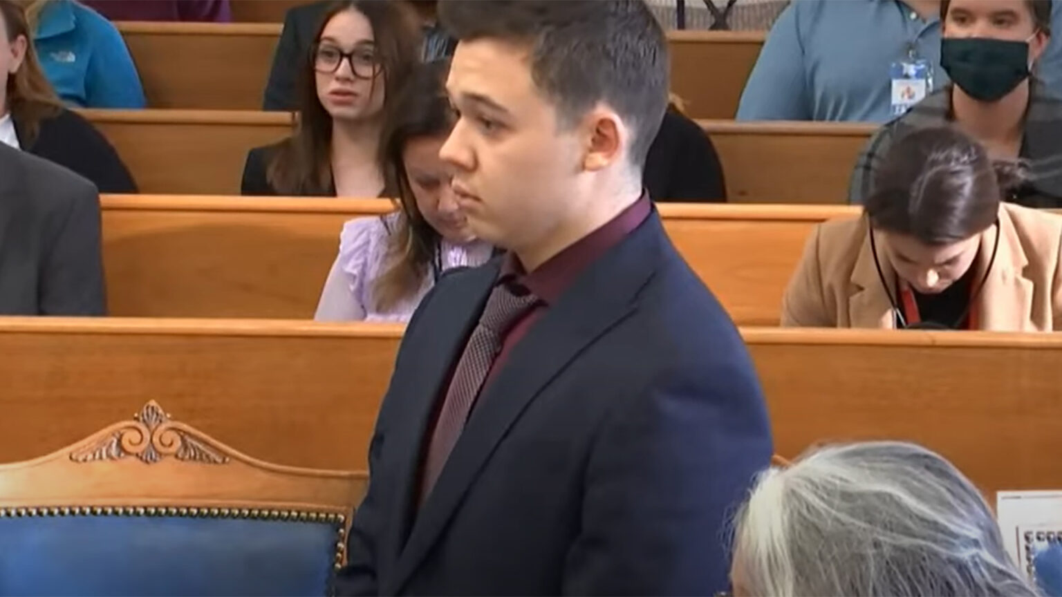 Kyle Rittenhouse stands in a courtroom with observers seated in the background.