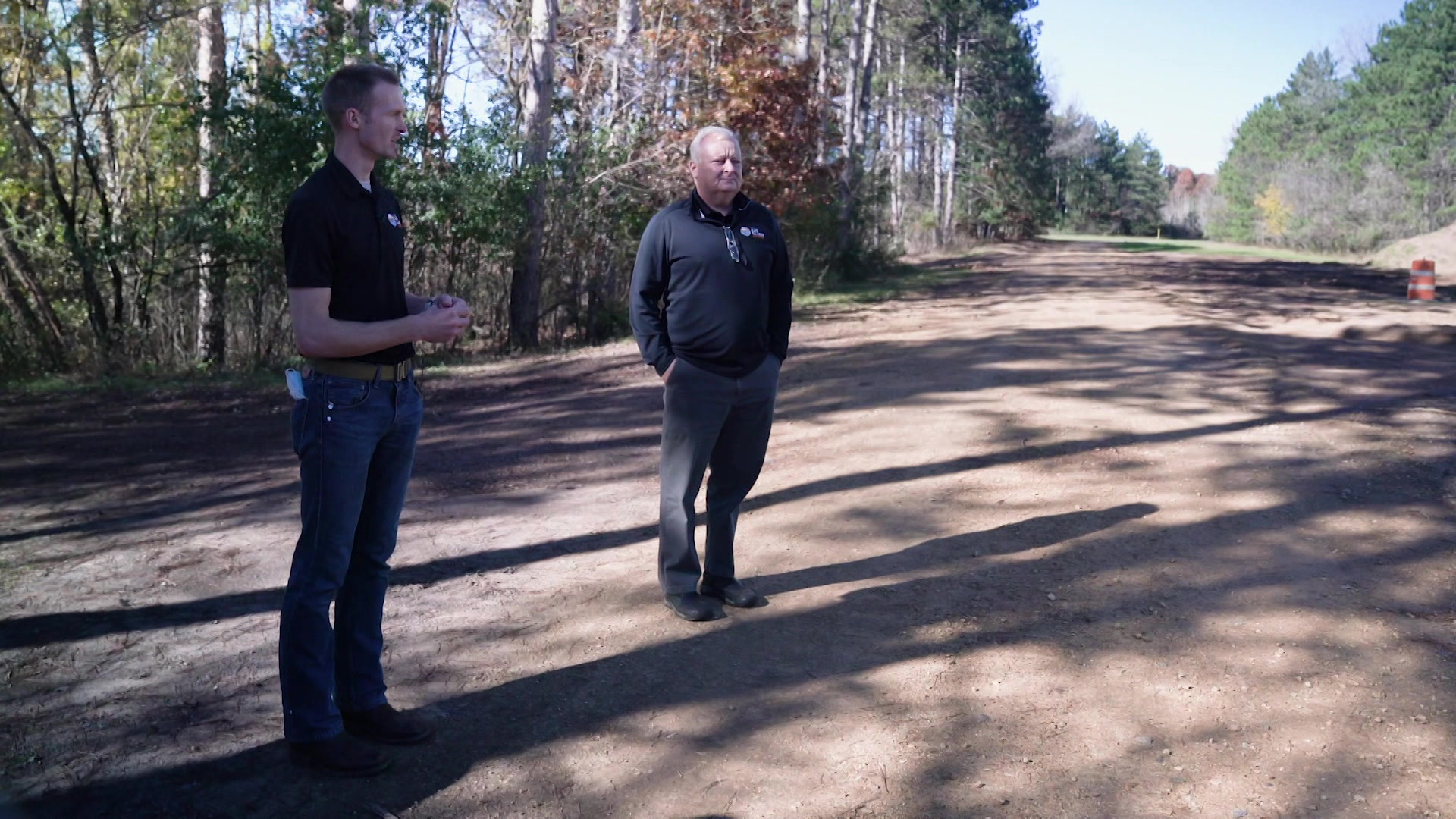 An Eau Claire utilities employee and Lane Berg stand on a field of bare earth with trees in the background.