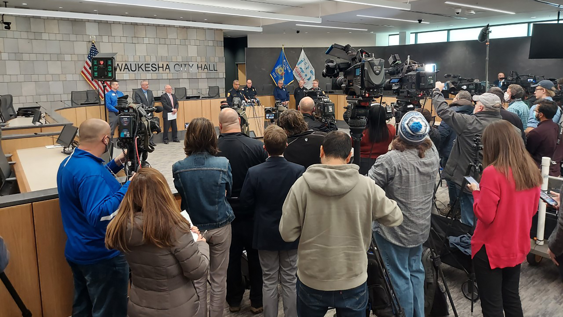 Reporters and TV cameras stand in a semicircle in a public meeting room facing local officials and law enforcement officers giving a press conference.