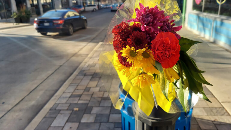 A bouquet of flowers is attached to a metal poll on a sidewalk with cars driving down the adjacent road.