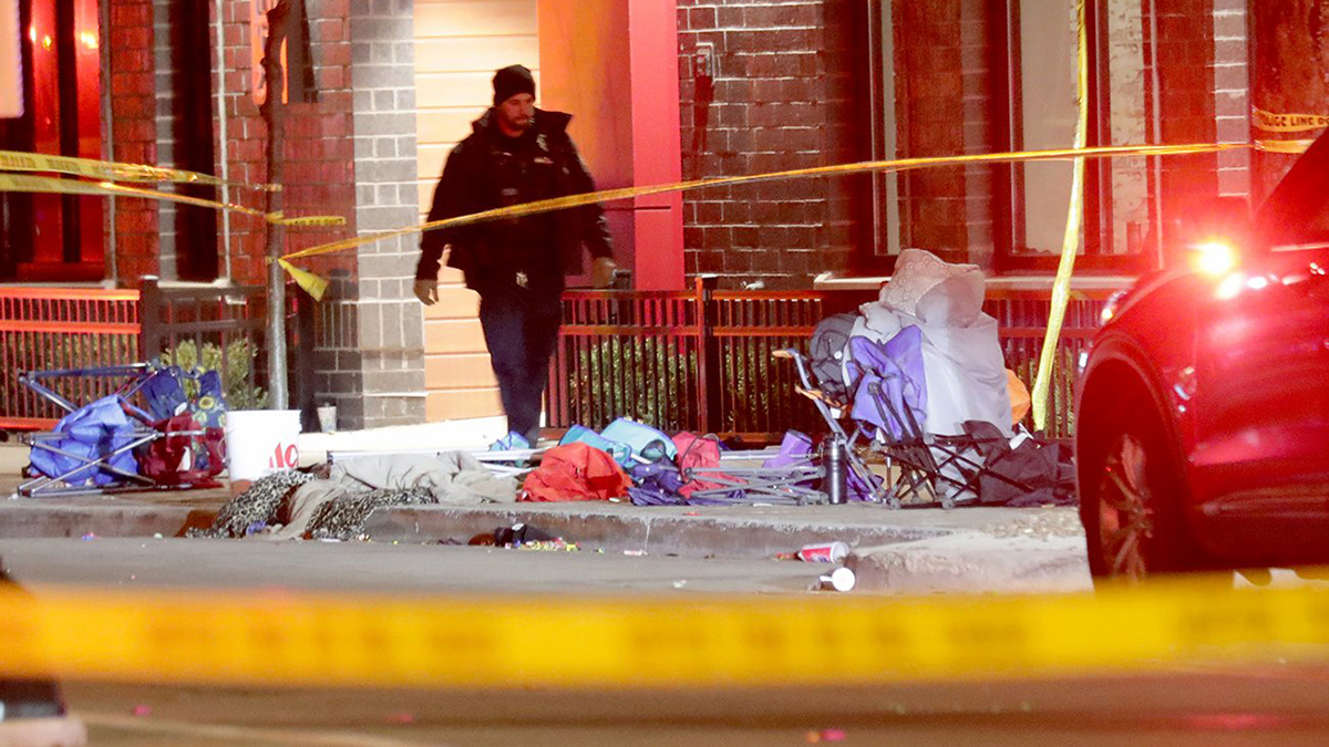 A police officer walks on a sidewalk strewn with abandoned chairs and personal items, with yellow police tape wrapped around trees.