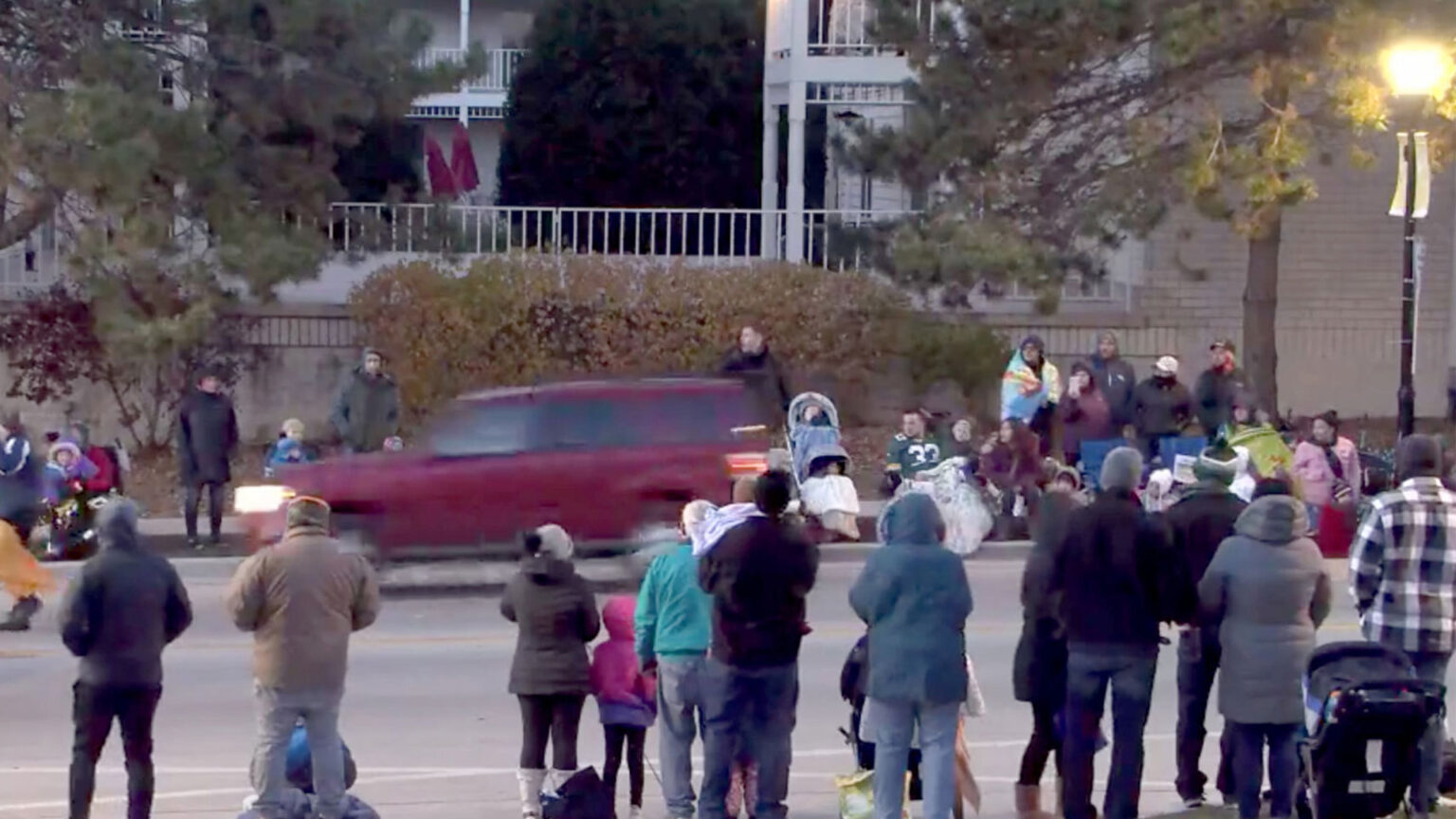 A blurry red SUV drives down a street with onlookers watching from the sidewalk.