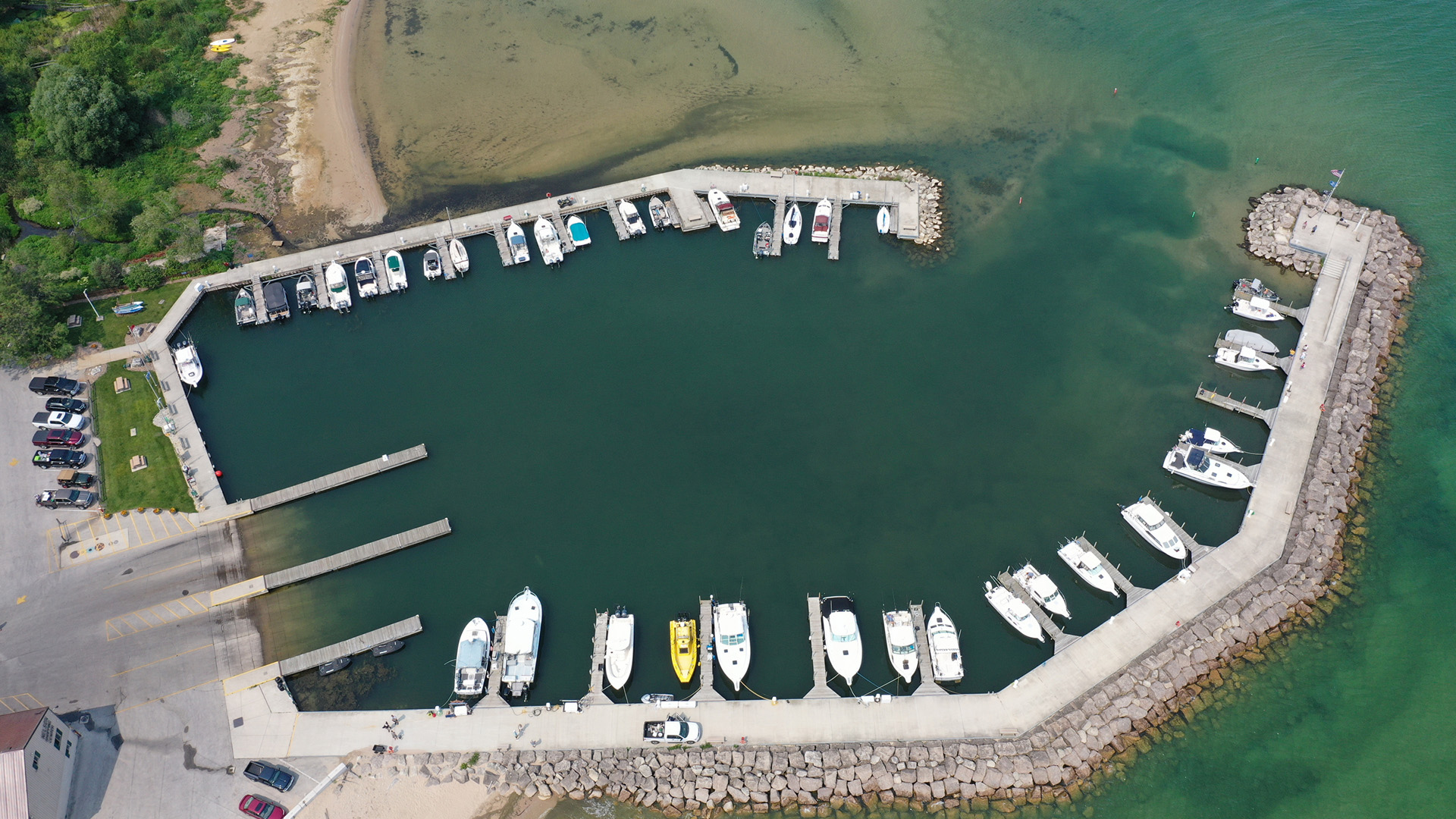 An aerial photo shows personal boats docked on piers on the inside of a ringed harbor surrounded by stacked boulders.