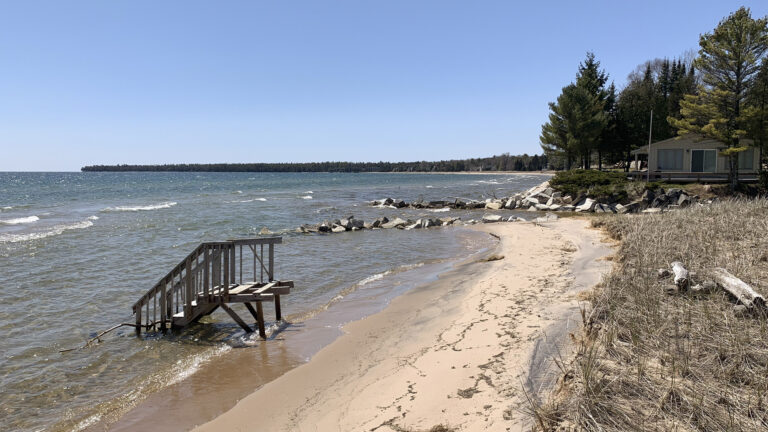 A wooden staircase stands alone on a sandy beach in the foreground with a house and wooded shoreline in the background.