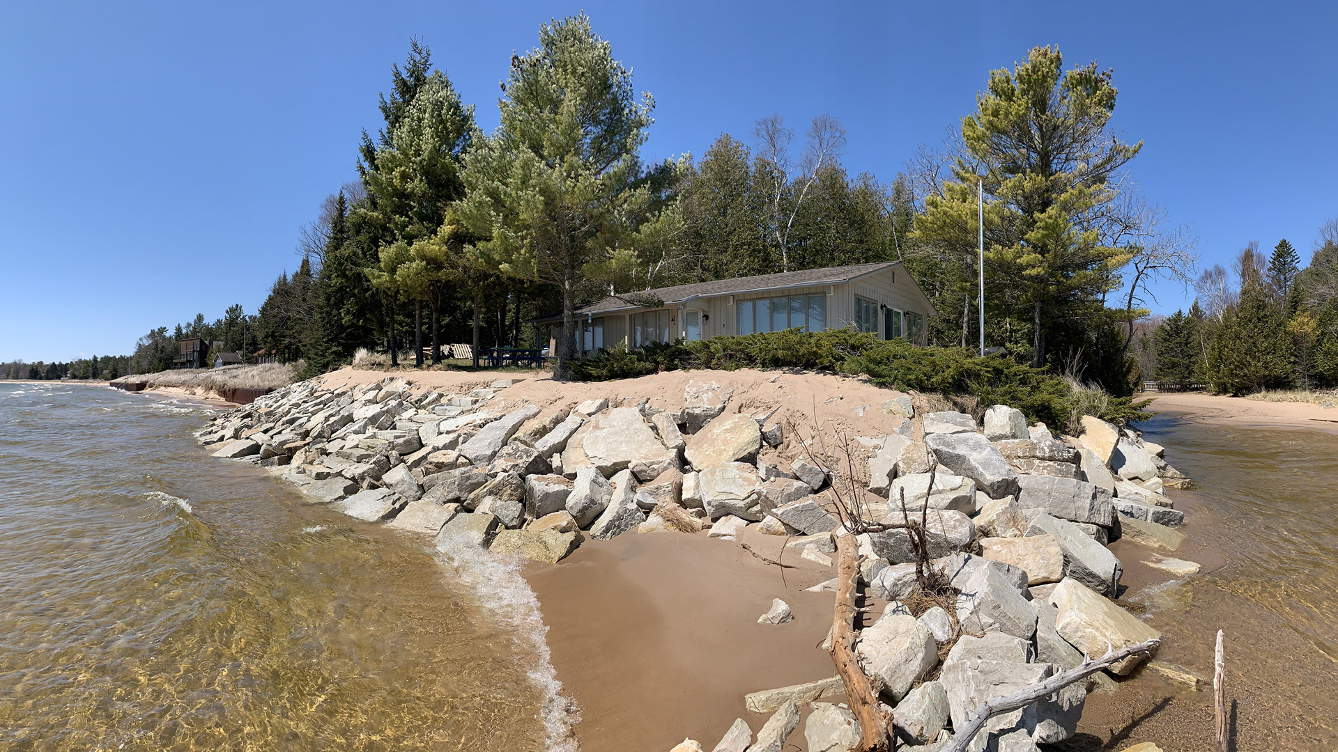 Boulders line a sandy shoreline in front of a house surrounded by trees.