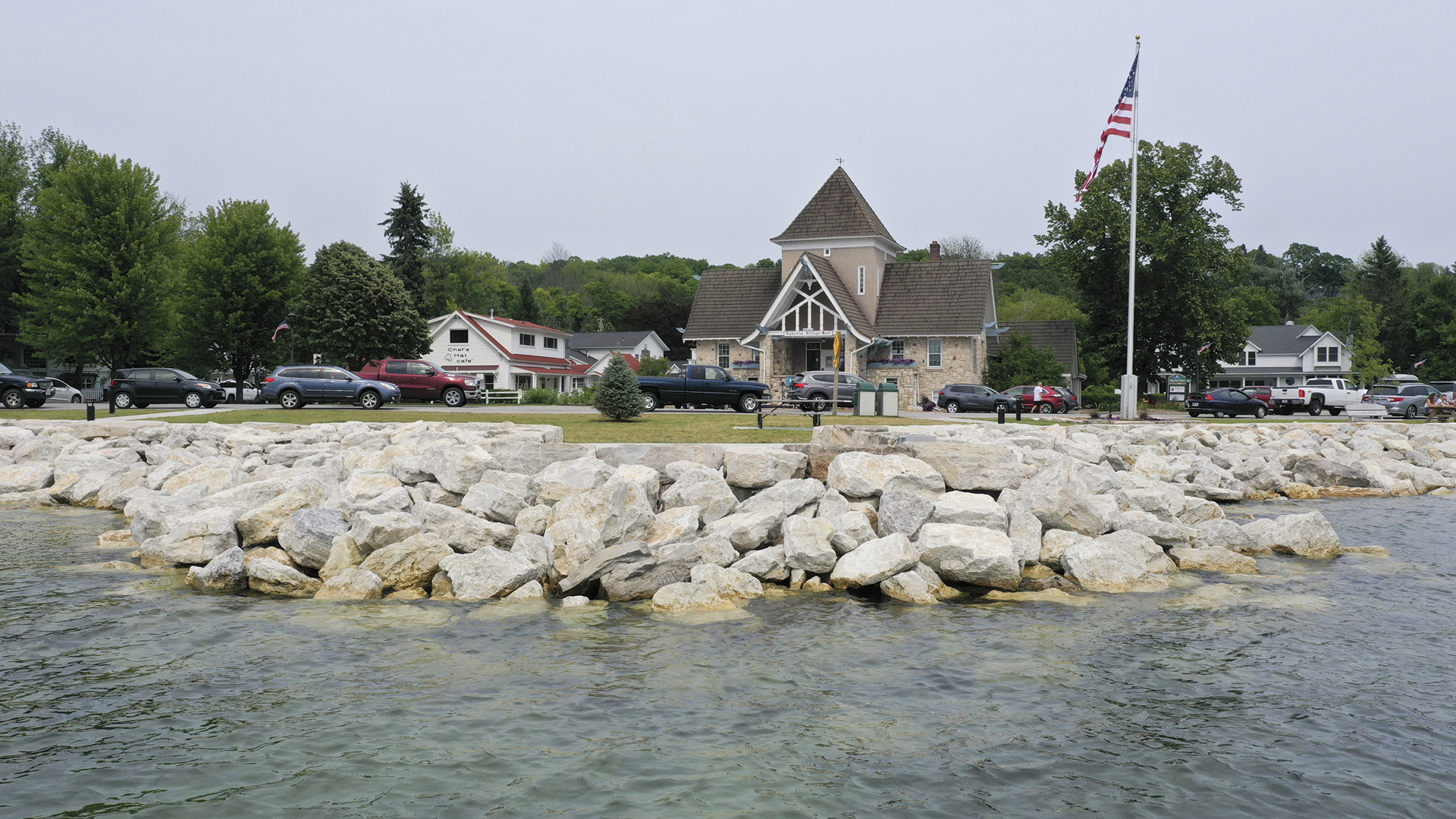 Large boulders line a lake shoreline with buildings and cars on the land above.