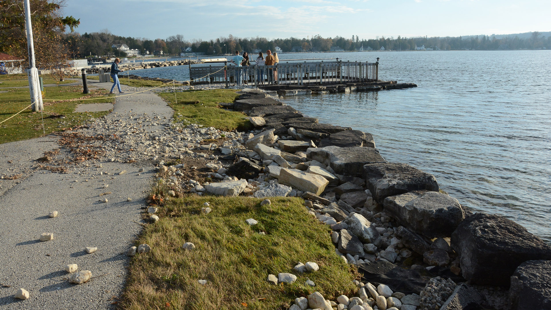 Pedestrians stand on a waterfront pier in the background with rocks strewn across a walkway and lawn in the foreground.